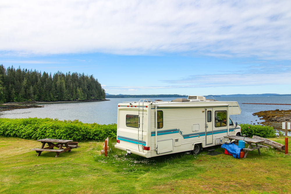 An RV on the shore of a lake surrounded by grass and tall trees with picnic tables next to it
