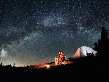 Two people sitting in front of a campfire by a tent at night with the sky full of stars while camping in Oklahoma