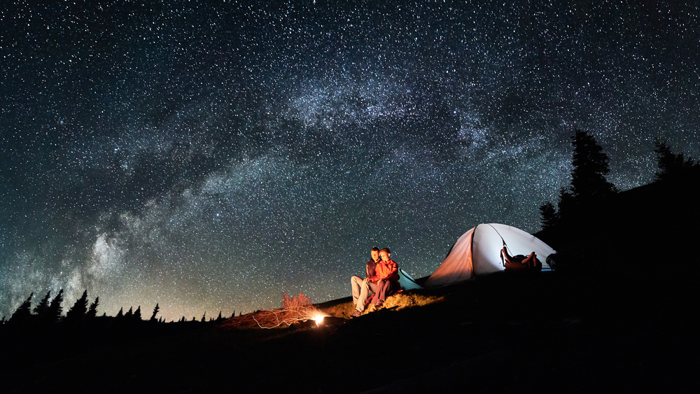 Two people sitting in front of a campfire by a tent at night with the sky full of stars while camping in Oklahoma