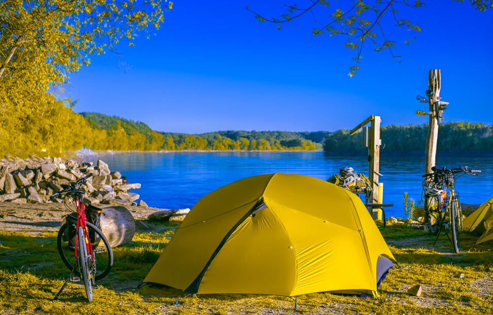 A greenish yellow tent on the side of a lake with bikes and electric hookup 