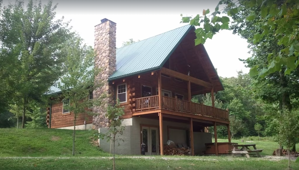 Large cabin with balcony surroud by trees. There is a table and hot tub inf ront of the cabin. 
