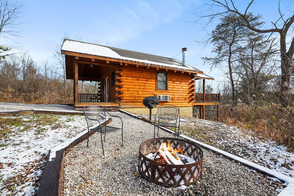 Cabin with hot tubs in the snow. the cabin has a balcony front and back. There is also a firepit and two chairs at the front of the picture.  