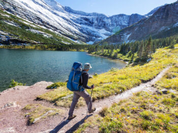 man walking with a daypack and a hiking stick on a trail with a lake and mountains hiking in montana