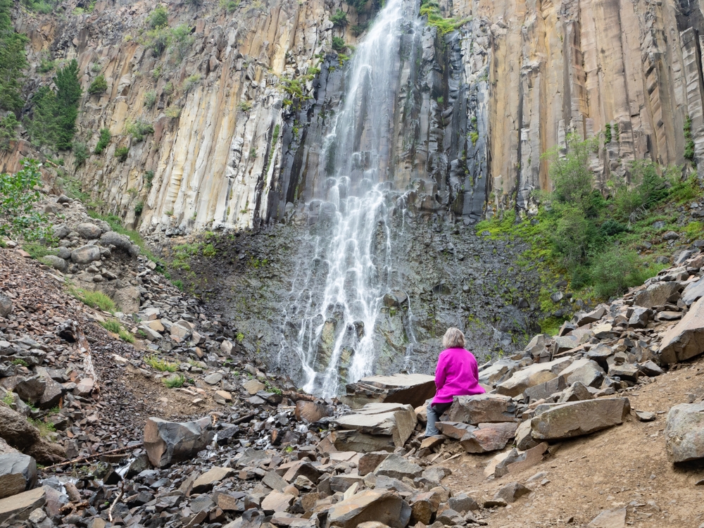 woman wearing a purple jacket sitting on rocks in front of a waterfall hiking in montana