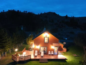 wooded cabin lit up with lights featuring white chairs on the deck surrounded by mountains