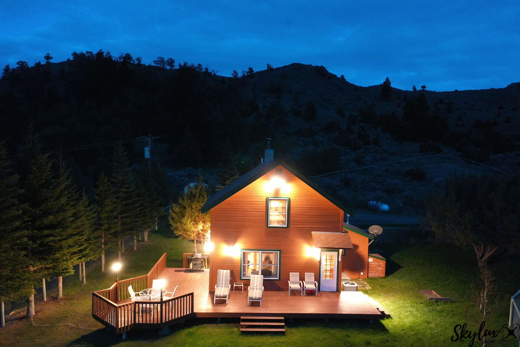 wooded cabin lit up with lights featuring white chairs on the deck surrounded by mountains