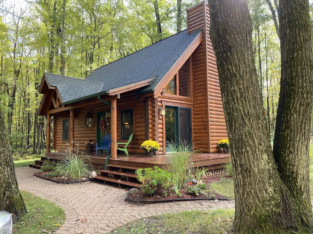 A log cabin with a porch and two chairs on it. There are woods behind the cabin and a paved area in front. 