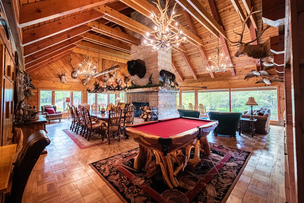The inside of a grand cabin with a high ceiling and large windows you can see the dining table some soafs and a games table. 