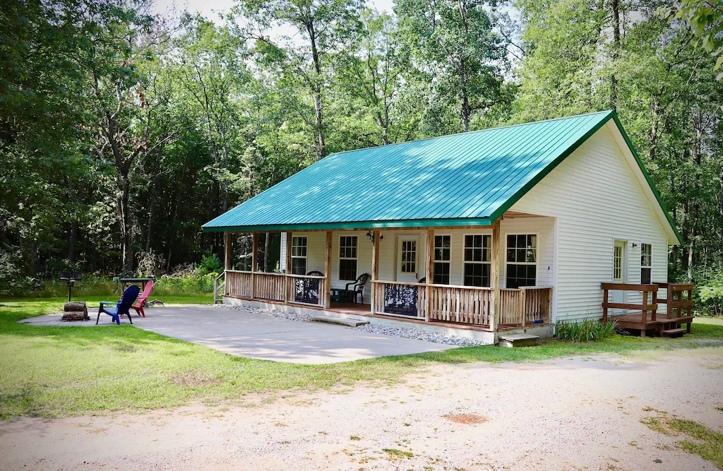 White cabin with a green woods in the woods. The cabin has a porch and there is a patio area.  