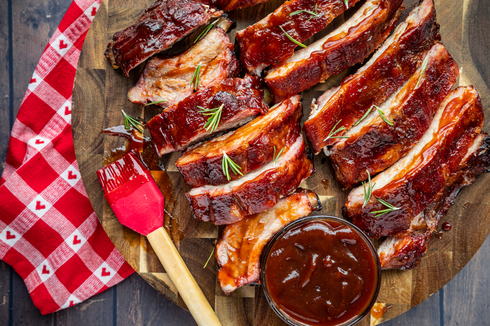 Barbeque ribs pieces and arranged on a round wooden cutting board with bowl of sauce and sauce brush and red and white checkered napkin with hearts on side