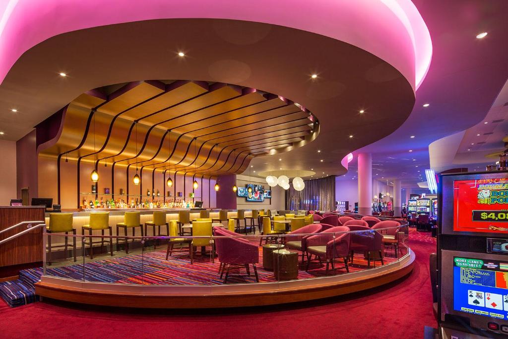 A bar area with pink and yellow chairs and pink light illuminating it. There are arcades off to the right. 