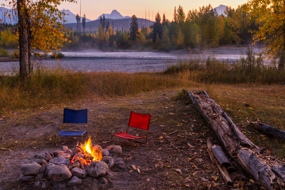 Sunset over a campsite with a fire and chairs next to a lake.