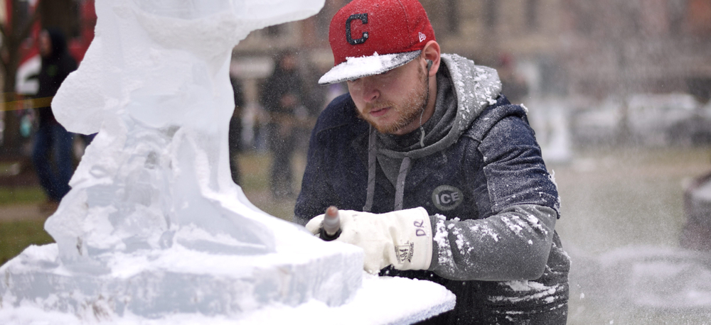 man bending down carving ice into a sculpture. He is wearing a read hat. 