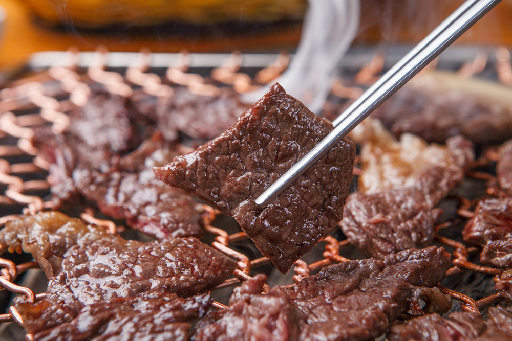 Close up of beef cooking on a grill. One of the pieces is been picked up on tongs. 