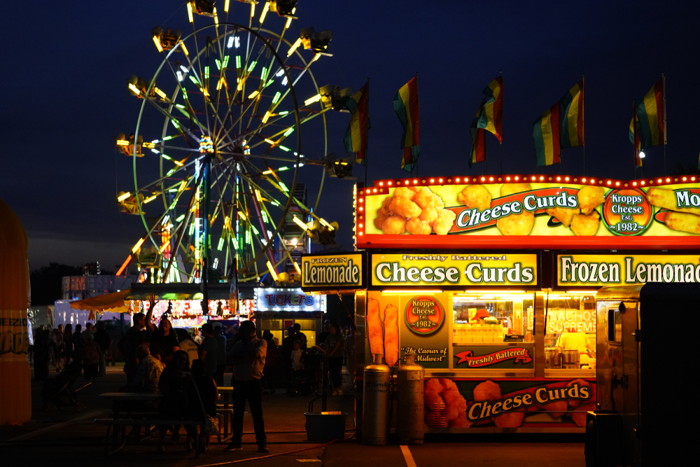 Nightime at the State Fair with a cheese curd stand in the foreground and a big wheel in the background.  