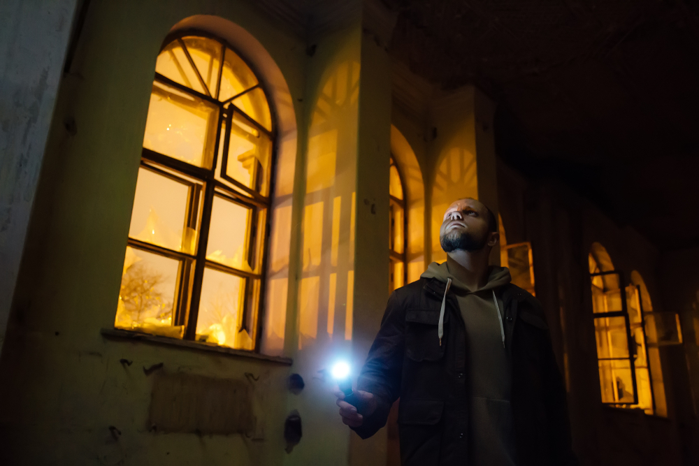 A man holding a flashlight in a dark building with street lights coming in through the windows