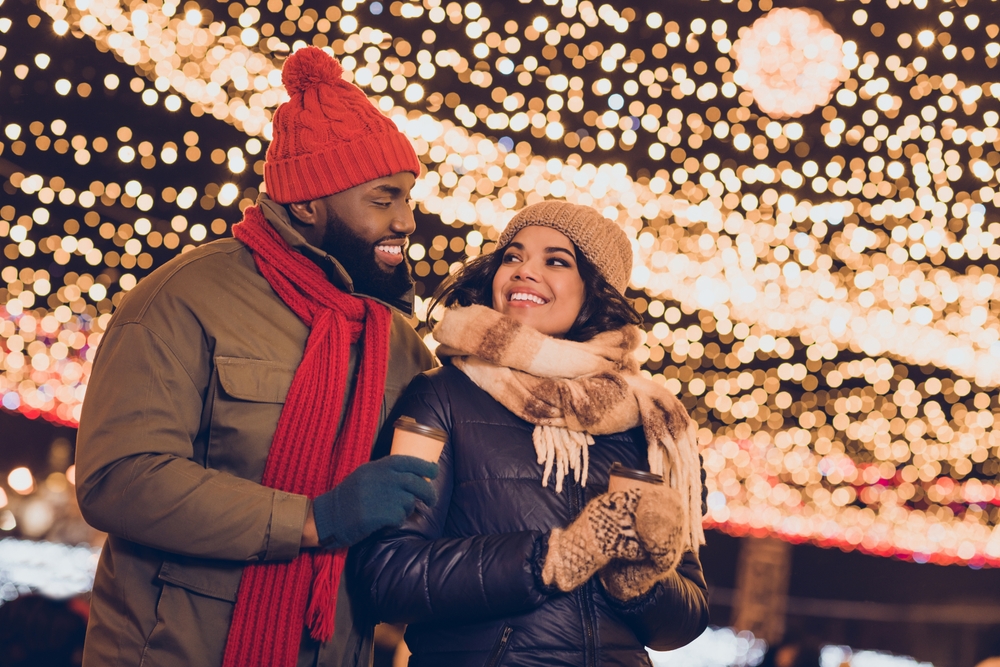 A couple outside under string lights during the winter looking at each other and holding warm drinks