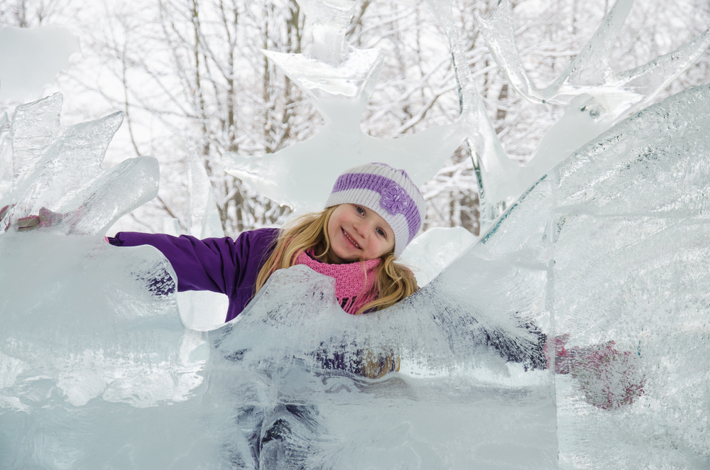 A young blonde girl posting behind a large ice sculpture in the winter