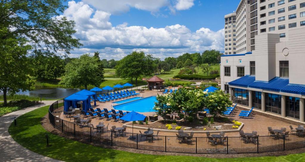 A white hotel by the lake with a swimming pool outside with blue deckchairs around.  