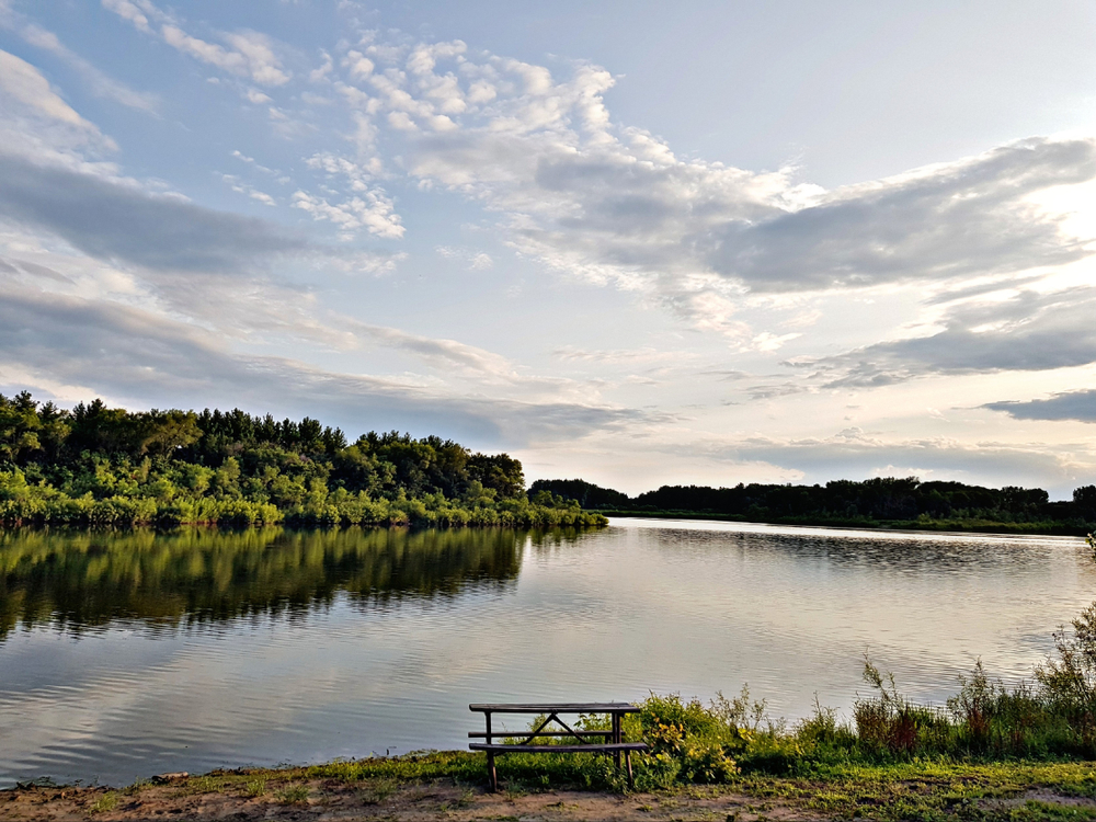 The view of a large lake with a picnic table on the shore and shores full of trees in the distance