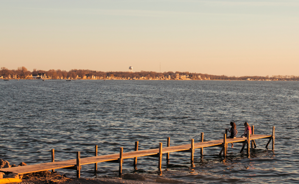 Two kids sitting on a dock in a lake with views of a city in the distance as the sun is setting