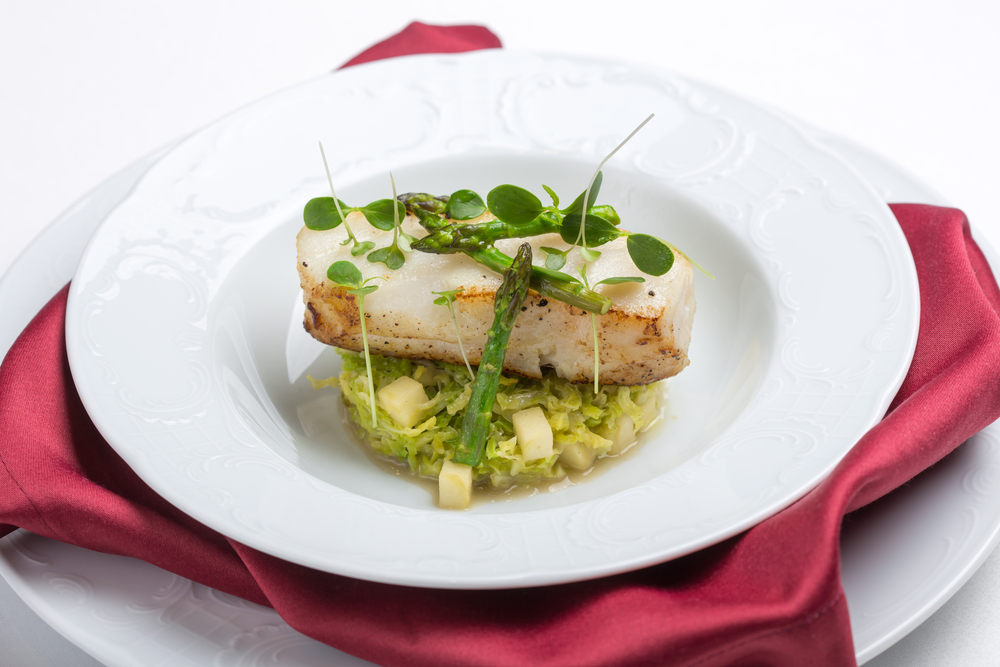 A fine dining dish of chilean sea bass with vegetables in a white bowl with a red cloth napkin under it