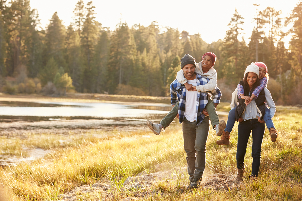 A family with the kids ridding piggy back on the parents walking on a trail next to a lake on a sunny day