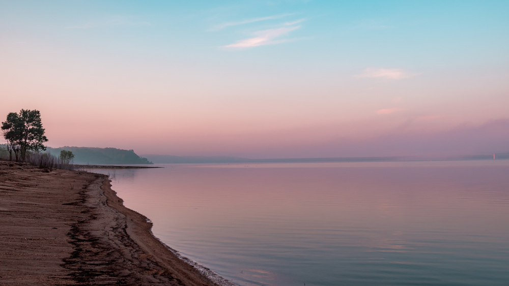 Looking down a sandy beach at one of the best lakes in Iowa as the sun is starting to rise, so the sky is pink, purple, and blue