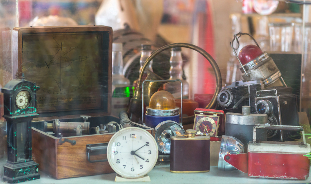 Assorted vintage/antique items, clocks, cameras, flasks, sextant, lamps behind shop window.