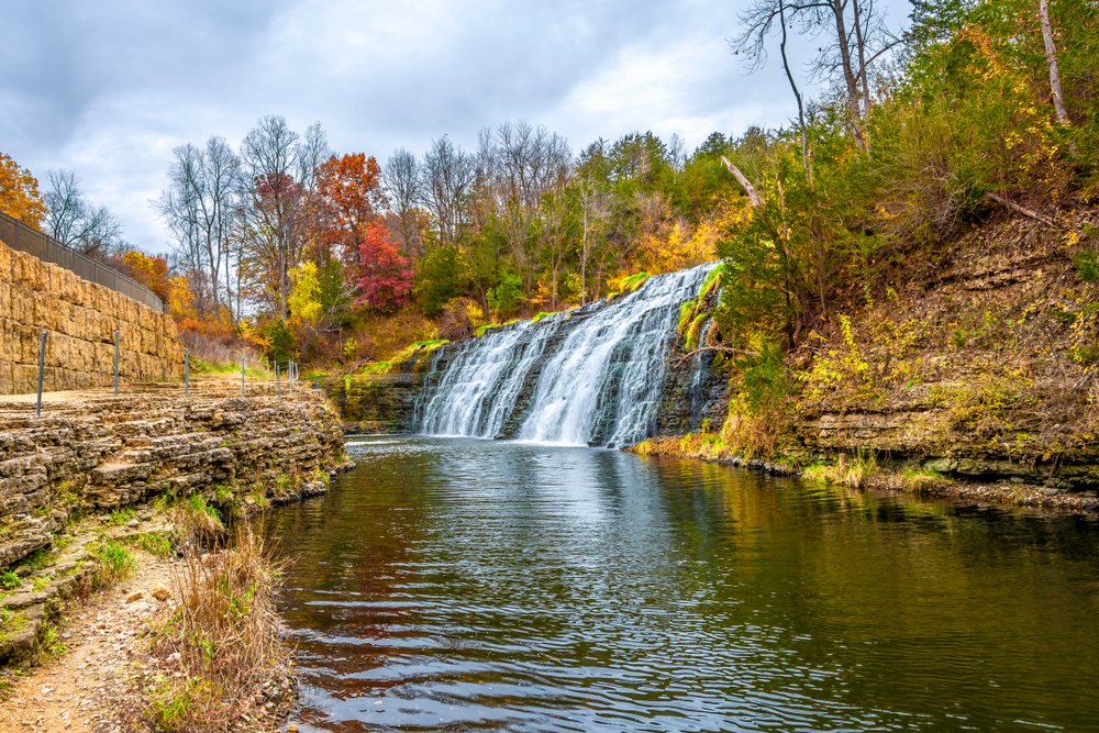 A cascading waterfall on the side of a hill surrounded by trees with fall foliage