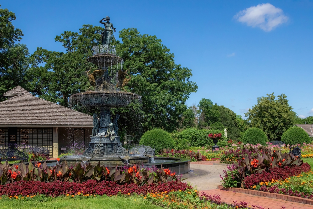 Tiered fountain with a statue on top surrounded by colorful flower beds in Clemens Gardens, one of the best things to do in St. Cloud, MN.