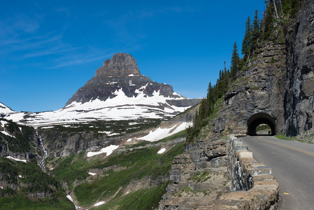 The Going-to-the-Sun Road hugging a rocky cliff and going through a tunnel with a view into a valley and a snowy mountain in the distance.