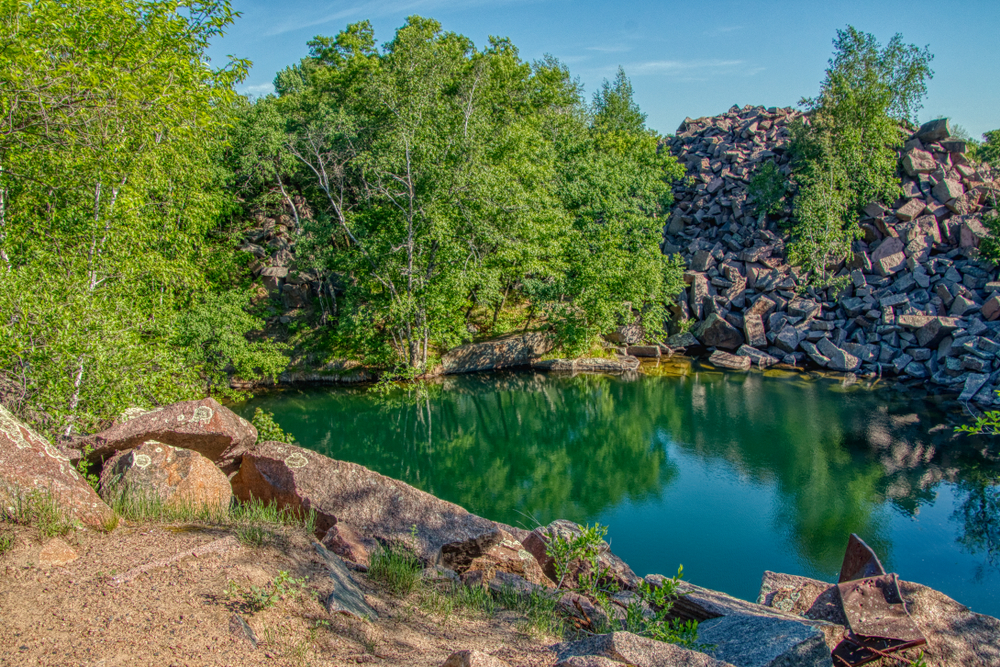 Bright blue water in a quarry surrounded with a pile of rocks and trees.