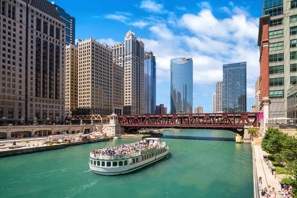 A cruise boats with lots of people on the upper deck going down the river among skyscrapers.