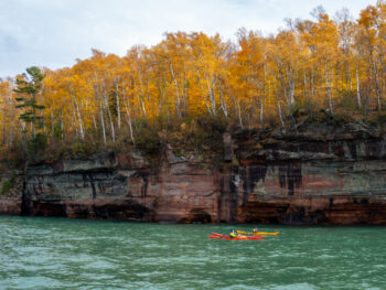 Two kayakers off the coast of the Apostle Islands which are covered in trees with yellow leaves, one of the best attractions in Wisconsin