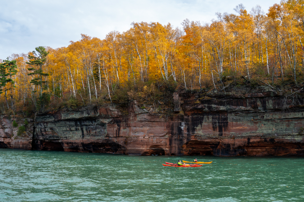 Two kayakers off the coast of the Apostle Islands which are covered in trees with yellow leaves, one of the best attractions in Wisconsin