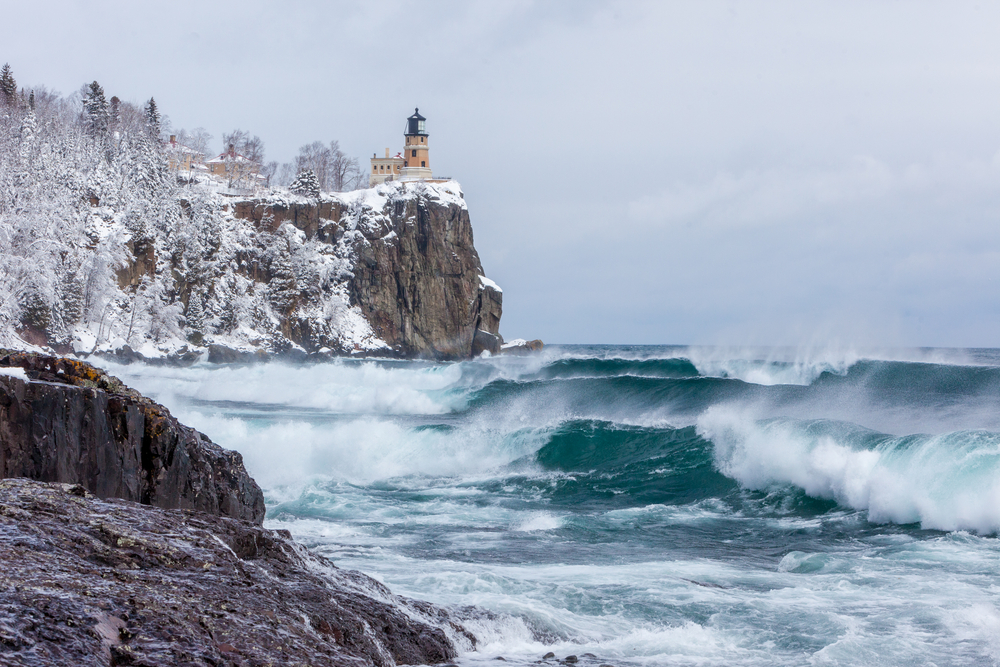 waves rolling onto the shoreline and a cliff with a lighthouse covered in snowy trees winter in minnesota