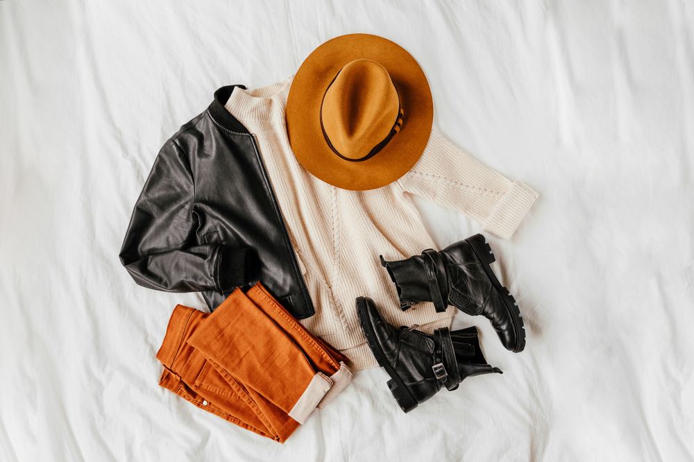 Flat lay image of a fall outfit including a beige sweater, black leather jacket, wide-brimmed hat, and black boots.