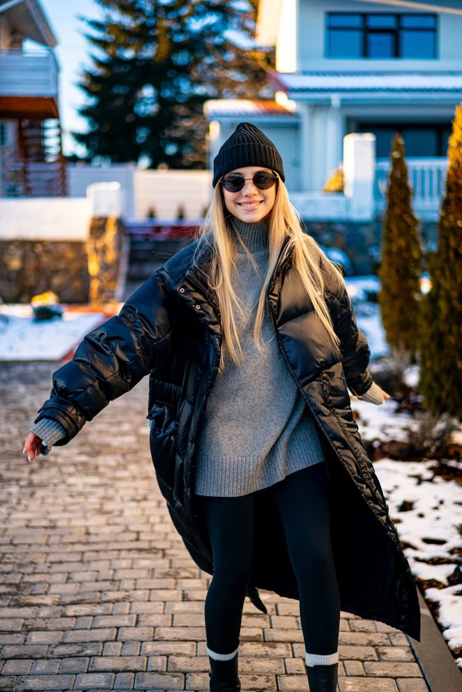 A young woman with long hair wears a warm black coat and beanie while standing on a stone pathway surrounded by snow.