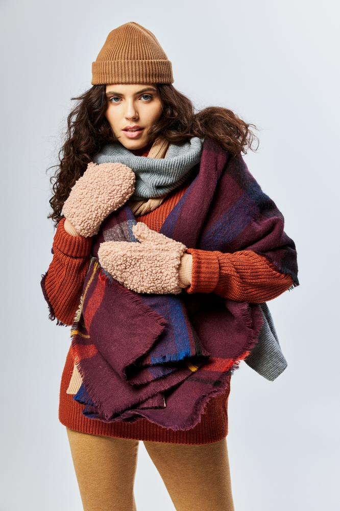 cozy layers, curly young woman in layered clothes with warm scarf and mittens on grey backdrop. Midwest winter packing guide.