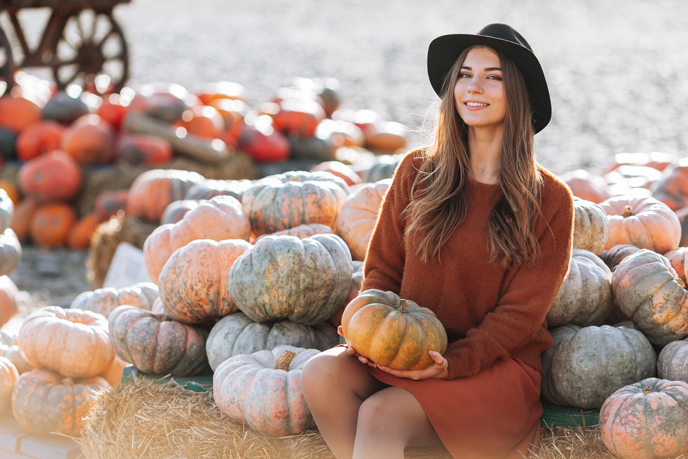 Woman sitting with pumpkin in hands near wagon with orange pumpkin on farmers market in brown sweater, dress and hat