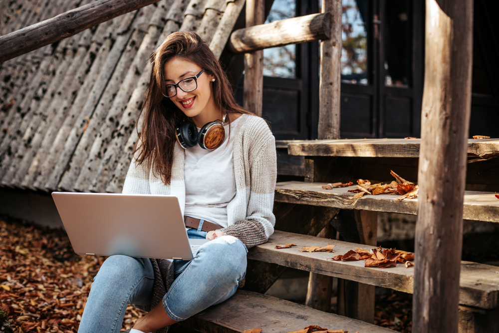 Woman sitting on wooden stairs in front of a cabin and working on a laptop computer