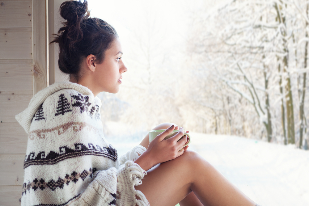 woman drinking cup of coffee wearing knitted nordic print poncho chilling out home by the window. Montana winter outfits.
