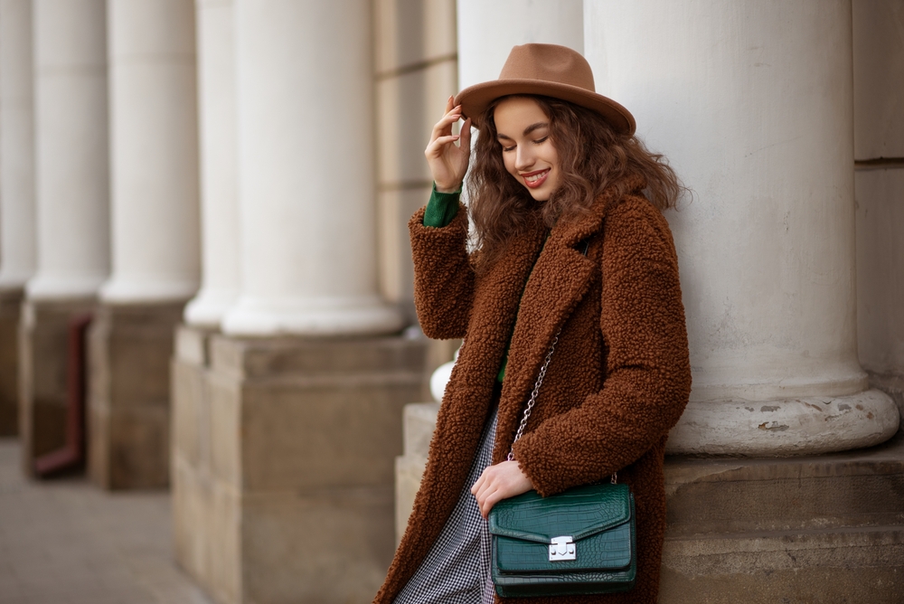 Woman wearing trendy brown faux fur teddy bear coat, with green textured leather shoulder bag, posing in street