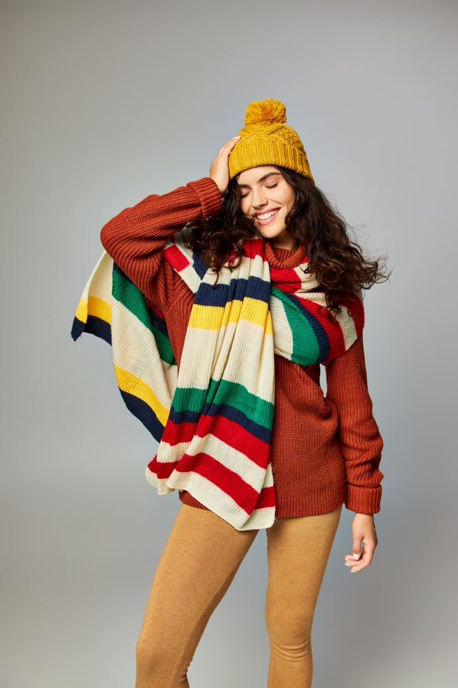 Woman in winter bobble hat and sweater with stripped scarf posing on grey background. Montana winter outfits.