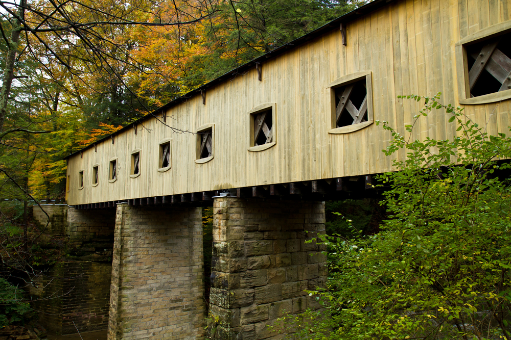 Close up picture of light brown covered bridge in Ashtabula County with stone columns surrounded by autumn colored greenery