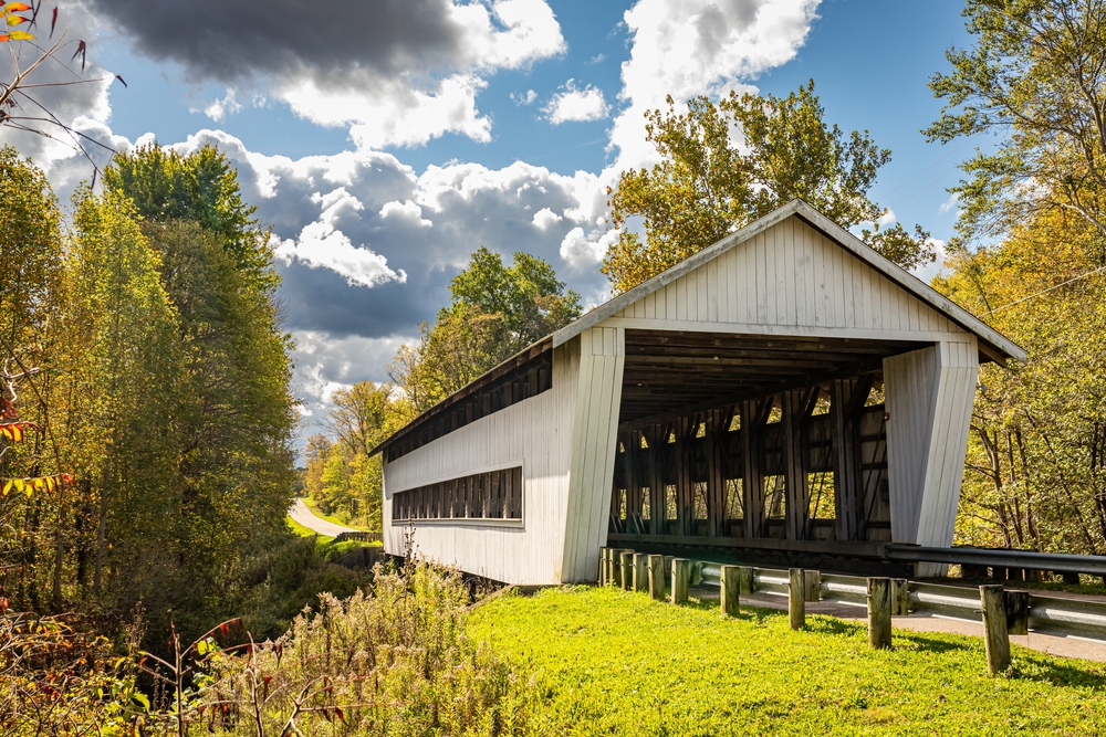 White covered bridge with road leading up to it with surrounded by greenery with chite clouds in blue sky.