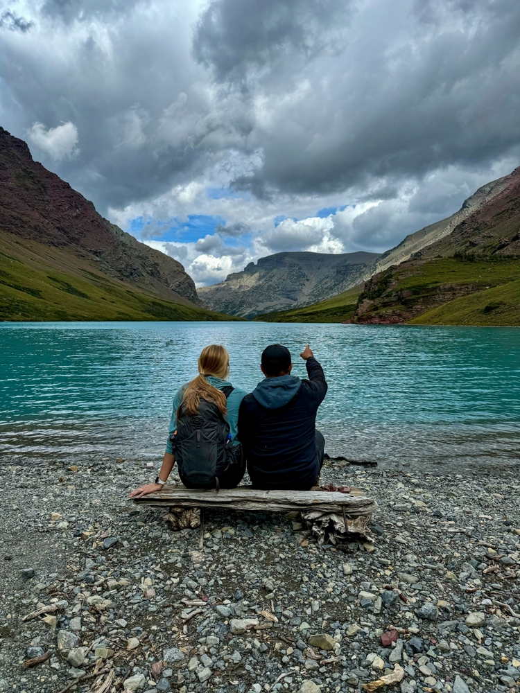 Man and woman sitting on log pointing and overlooking a blue lake with Mountain View. The article is about Romantic Montana getaways.