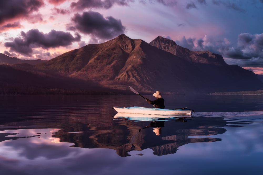 Adventurous Man Kayaking in Lake McDonald with American Rocky Mountains in the background. Colorful Sunset Sky. 
