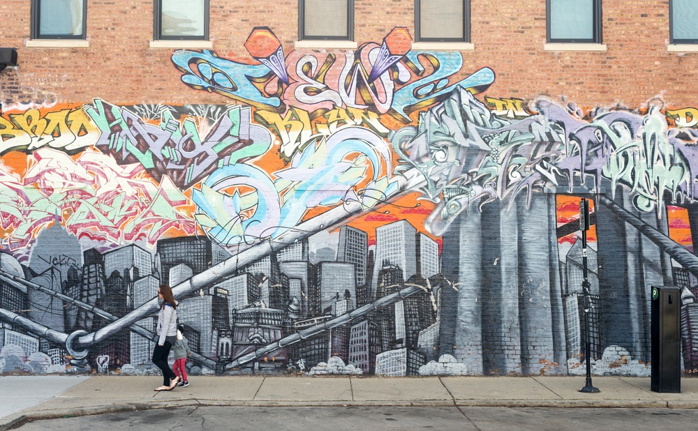 A mural on a red brick building with silver buildings and colorful writing 